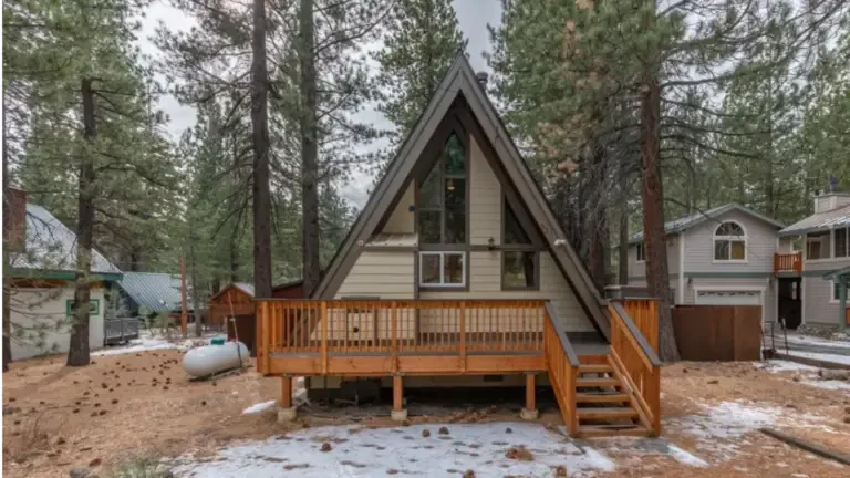 A-frame cabin surrounded by trees, featuring a wooden deck and stairs, with light snow on the ground. Nearby are other houses and a propane tank.