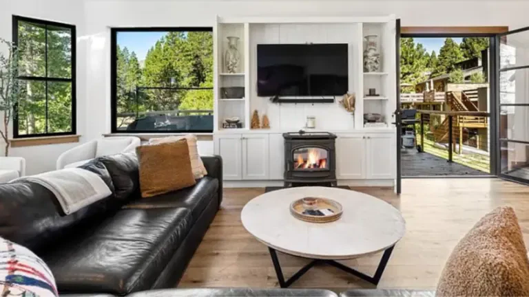 A cozy living room with a black leather sofa, a round coffee table, a fireplace, and a mounted TV. Large windows and open doors lead to a wooden deck with outdoor seating.
