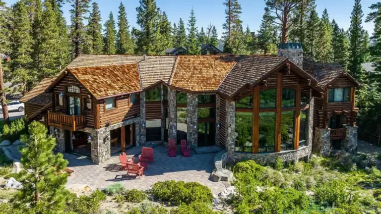 A large log cabin with stone features, surrounded by trees. It has multiple large windows, a slanted roof, and a patio with red chairs.