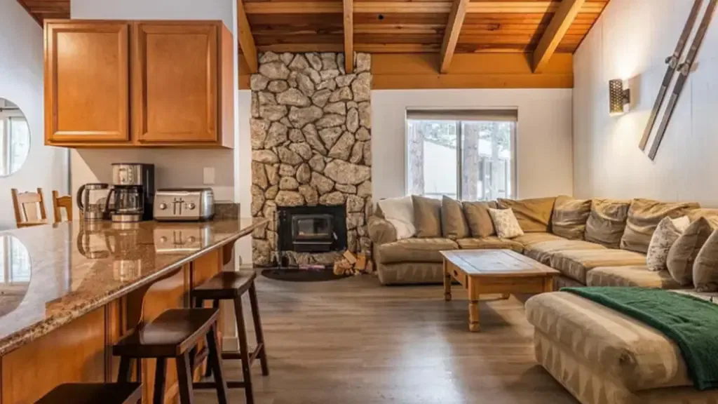 Cozy living room with a stone fireplace, wooden beams, and a large sectional sofa. A kitchen counter with stools and appliances is visible on the left. Natural light filters through a window.