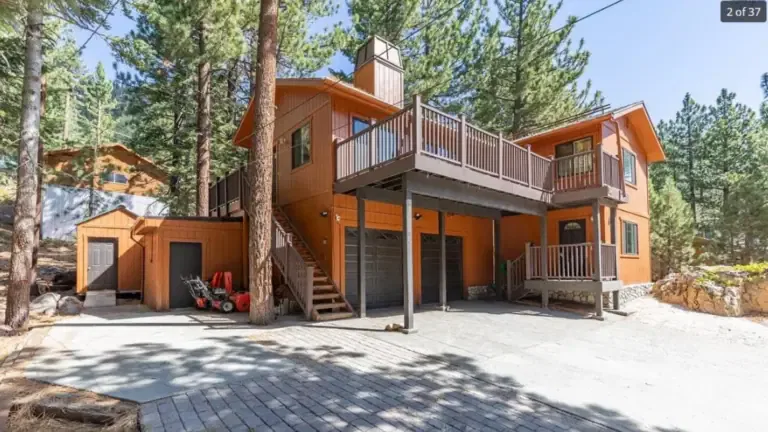 Two-story wooden cabin with a balcony, two garage doors, and a paved driveway, surrounded by tall trees.