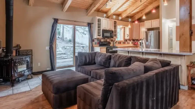 A cozy living room with a dark sectional sofa, wood-burning stove, and open kitchen. Wooden ceiling beams and sliding doors lead to a stone patio.