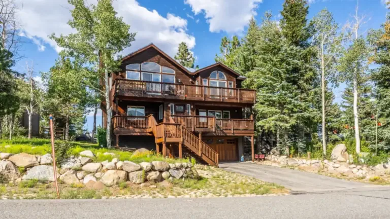 A large wooden cabin with multiple balconies is surrounded by trees and set on a stone foundation. A paved driveway leads to the entrance.