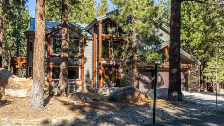 A modern, two-story cabin in a wooded area with large windows, stone accents, and a garage, surrounded by tall pine trees and rocks.