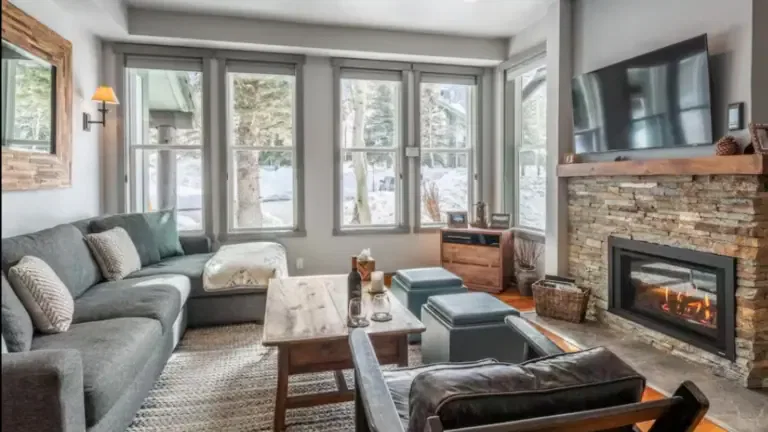 A cozy living room with a large sectional sofa, a wooden coffee table, a stone fireplace, and a wall-mounted TV. Tall windows show a snowy outdoor scene.