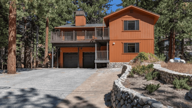 Two-story wooden house with a stone foundation, double garage, and balcony, surrounded by pine trees and a stone-lined driveway.