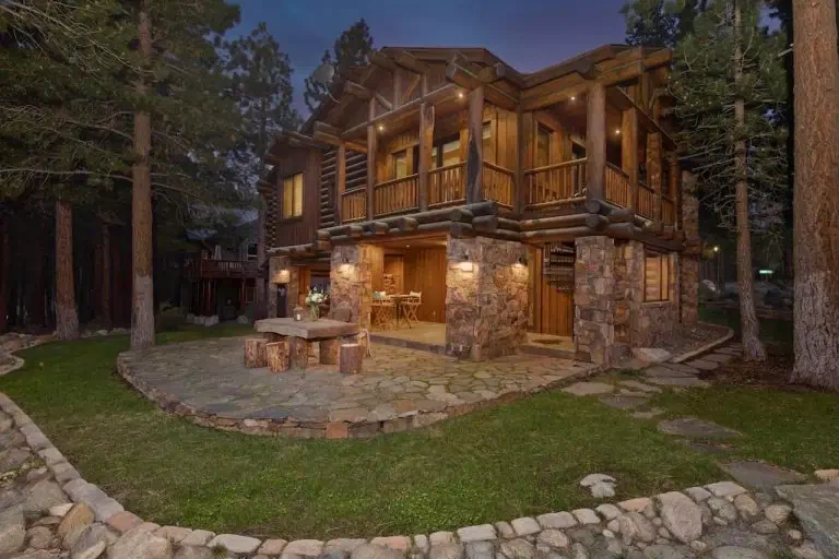 Two-story log cabin with stone base, outdoor stone patio, and rustic wooden furniture, surrounded by tall pine trees at dusk.