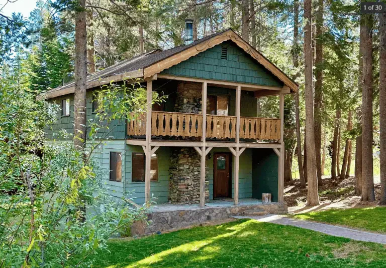 Two-story green wooden cabin with a stone chimney, balcony, and covered porch, surrounded by tall trees and grass in a forested area.