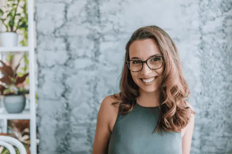 A woman with long brown hair and glasses smiles at the camera, standing in front of a textured light gray wall with some plants visible on the left.