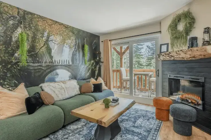 A cozy living room with a green sofa, wooden table, fireplace, wall mural of a forest scene with a bridge, and sliding doors leading to a balcony with trees outside.