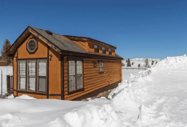 A small wooden cabin with multiple windows sits surrounded by deep snow under a clear blue sky in a rural winter landscape.