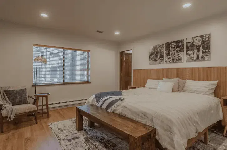 A cozy bedroom with a large bed, wooden bench, armchair, window with blinds, and three dog-themed pictures on the wall above the bed.