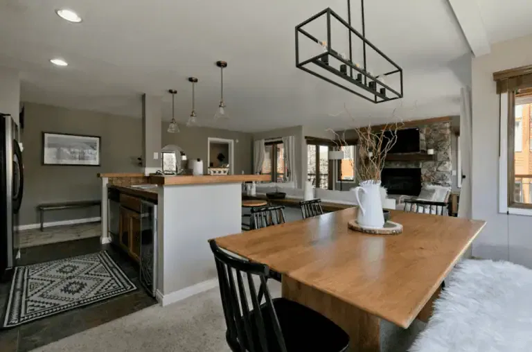 A modern dining area with a wooden table and black chairs is next to an open kitchen and living room with a stone fireplace and large windows.
