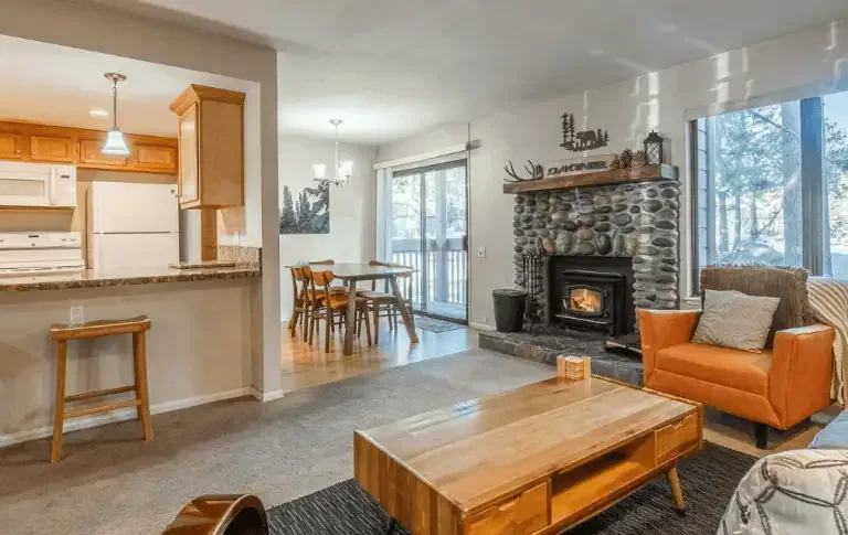 Open living space with a stone fireplace, orange armchair, wooden coffee table, and dining area next to sliding glass doors leading outside.