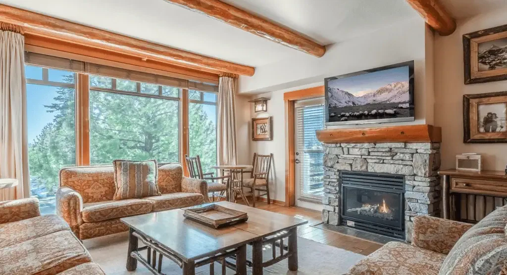 A cozy living room with patterned sofas, a wooden coffee table, a stone fireplace, and a TV above the mantel, featuring large windows with a view of pine trees.