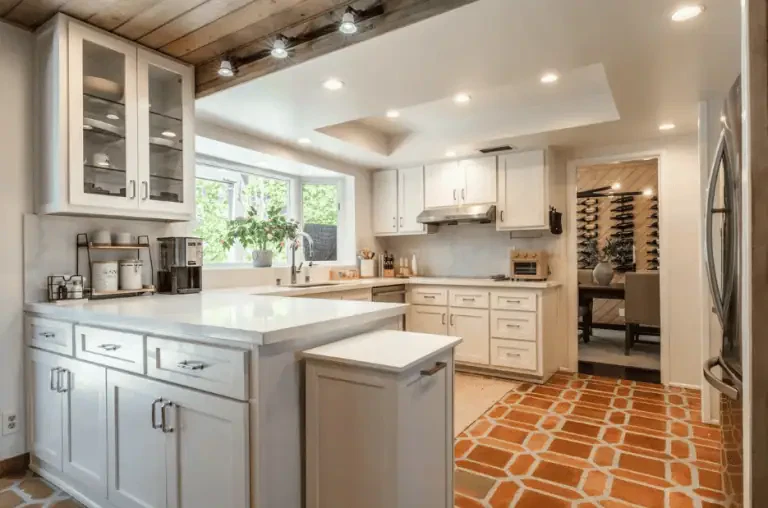 Bright kitchen with white cabinets, marble countertops, built-in appliances, a large window, and terracotta tile flooring. Dining area with a wine rack visible through an open doorway.