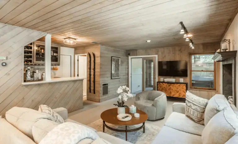 A cozy living room with wood-paneled walls and ceiling, a round coffee table, upholstered seating, a wall-mounted TV, a fireplace, and a small bar area with shelves and wine storage.