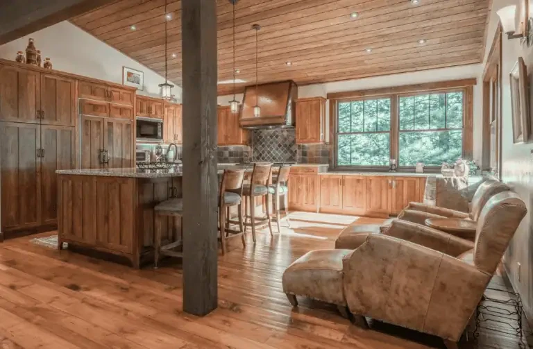 Spacious kitchen with wooden cabinets, central island with bar stools, leather armchairs, large window, and wood-paneled floor and ceiling.