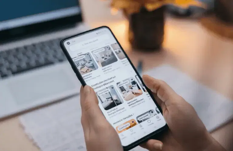 A person holds a smartphone displaying a property listings app, with images and prices of apartments, in front of a laptop and notepad on a desk.