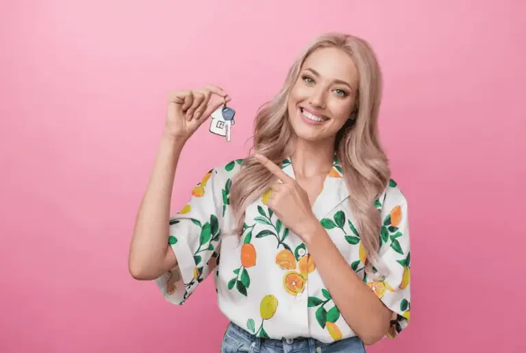 A smiling woman with long blonde hair holds and points to a house-shaped keychain labeled 