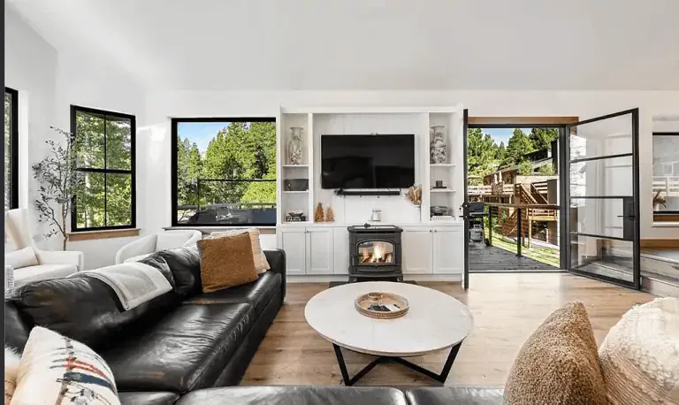 Modern living room with black leather sofa, round coffee table, fireplace, built-in shelves, wall-mounted TV, large windows, and open doors leading to an outdoor deck with forest views.
