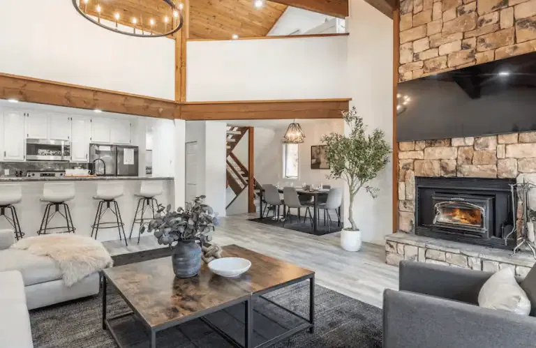 Modern open-concept living space with stone fireplace, large TV, kitchen with bar stools, dining area, and wood accents throughout.