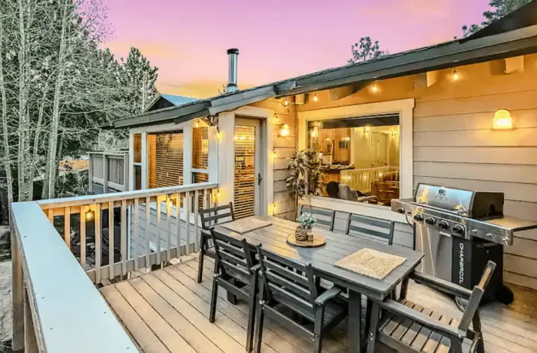 Wooden deck with outdoor dining table, chairs, and a gas grill attached to a house, lit by string lights at dusk; large window shows the interior.