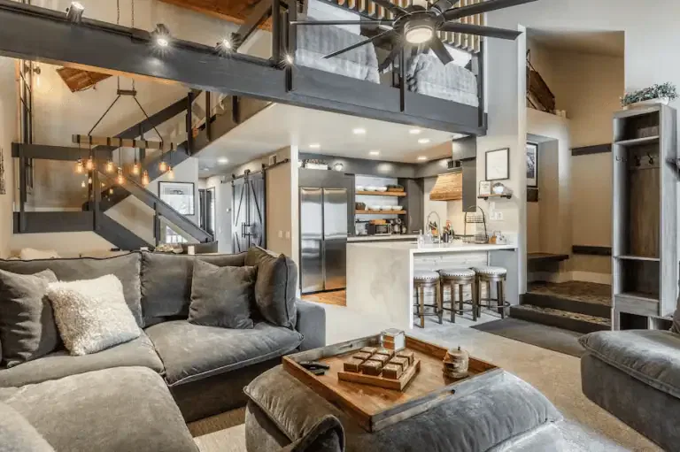 Modern open-plan living area with grey sectional sofa, wooden coffee table, kitchen with bar stools, lofted beds, and exposed beams. Neutral tones and natural light fill the space.