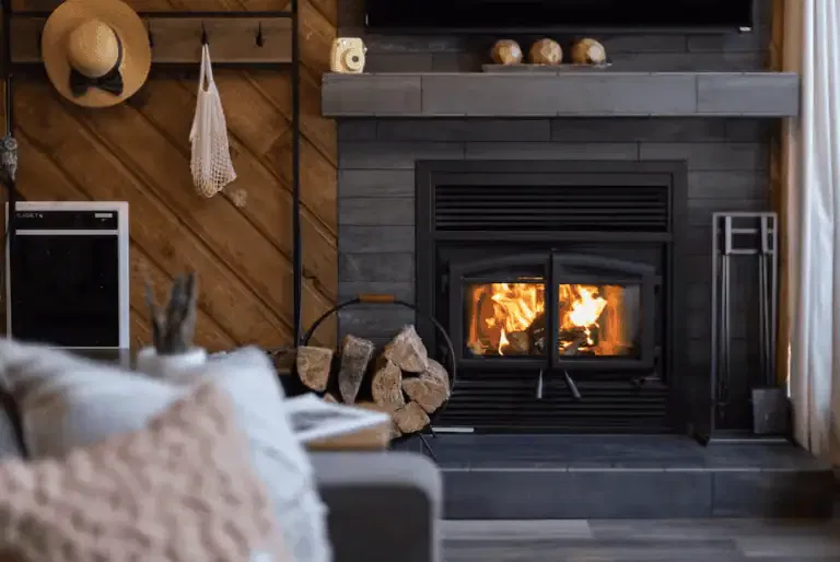 A modern living room with a lit fireplace, stacked firewood, a gray sofa, and a wooden accent wall with hanging items.