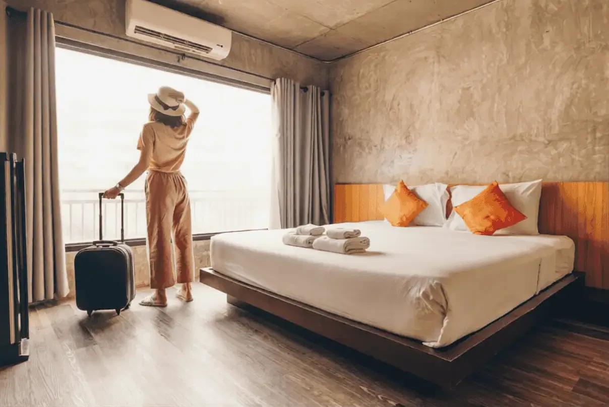 Let's talk A person with a suitcase stands by a large window in a modern hotel room with a double bed, white linens, and two orange pillows.