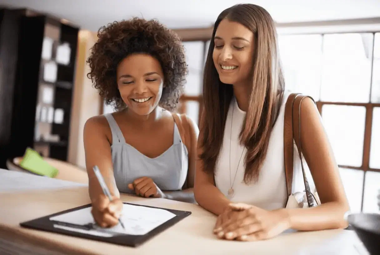 Let's talk Two women stand at a counter; one is smiling while signing a document on a clipboard, and the other looks on, also smiling.