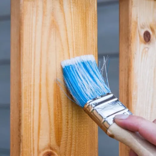 A person is painting a wooden fence with a paint brush.