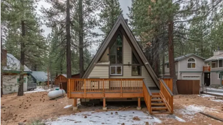 A-frame cabin surrounded by trees, featuring a wooden deck and stairs, with light snow on the ground. Nearby are other houses and a propane tank.