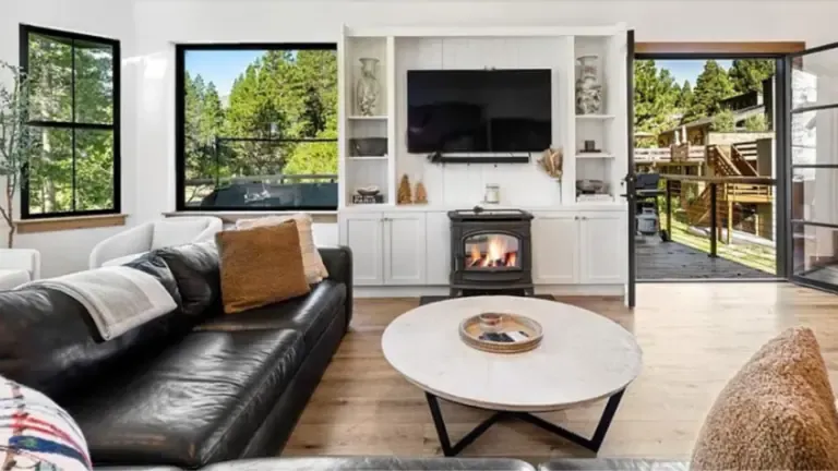 A cozy living room with a black leather sofa, a round coffee table, a fireplace, and a mounted TV. Large windows and open doors lead to a wooden deck with outdoor seating.