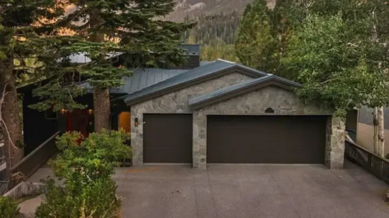 A stone house with a dark roof and two garage doors is surrounded by trees.