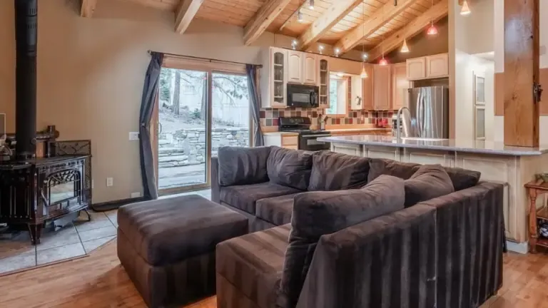 A cozy living room with a dark sectional sofa, wood-burning stove, and open kitchen. Wooden ceiling beams and sliding doors lead to a stone patio.