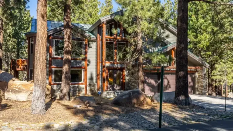 A modern, two-story cabin in a wooded area with large windows, stone accents, and a garage, surrounded by tall pine trees and rocks.
