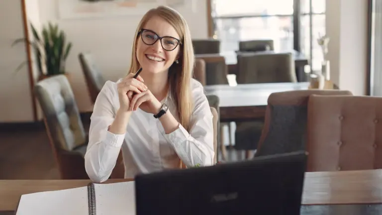 A person with glasses smiles while sitting at a table with a laptop and notebook in a bright room with large windows and chairs in the background.