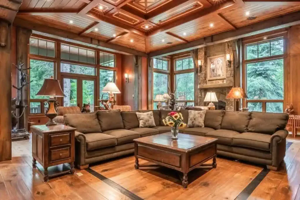 Spacious living room with brown sectional sofa, wooden coffee table, large windows, stone fireplace, wood-paneled ceiling, and forest view outside.