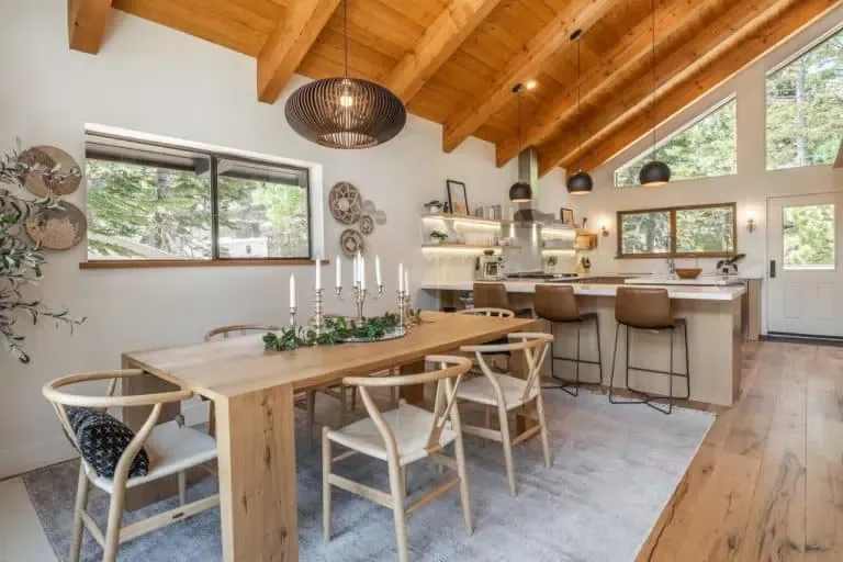 A modern, open kitchen and dining area with wooden ceiling and floors, a large dining table with chairs, barstools at an island, and large windows letting in natural light.