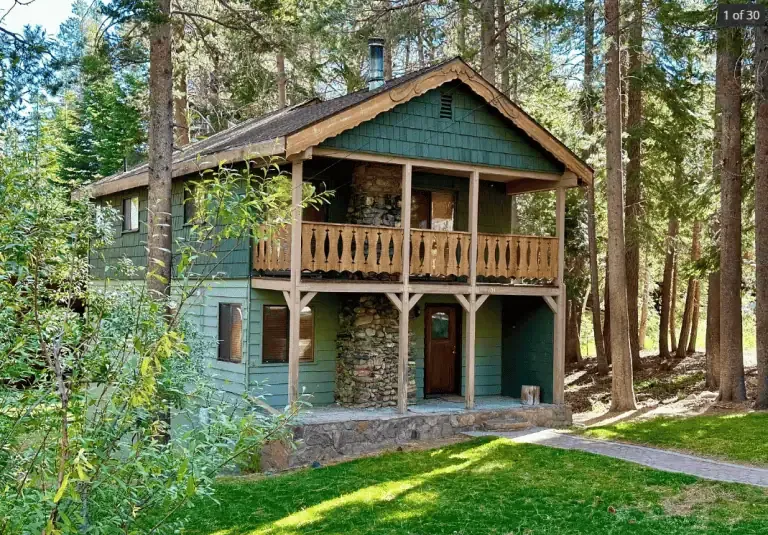 Two-story green wooden cabin with a stone chimney, balcony, and covered porch, surrounded by tall trees and grass in a forested area.