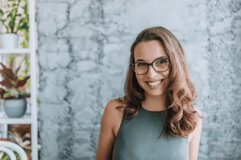 A woman with long brown hair and glasses smiles at the camera, standing in front of a textured light gray wall with some plants visible on the left.