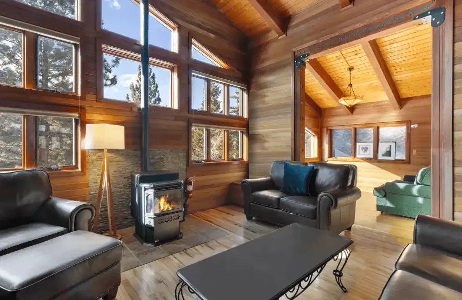 A cozy living room with leather sofas, a wood-burning stove, large windows, wood-paneled walls, and a view into an adjoining room with a green sofa and chandelier.