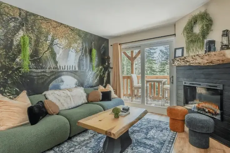 A cozy living room with a green sofa, wooden table, fireplace, wall mural of a forest scene with a bridge, and sliding doors leading to a balcony with trees outside.