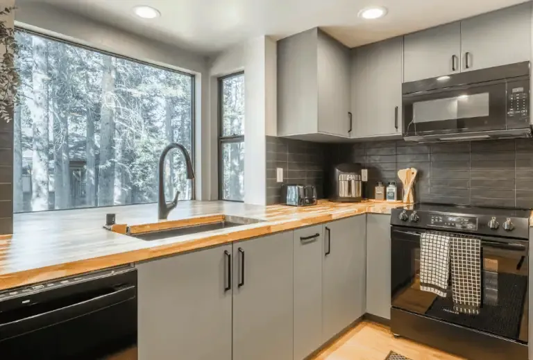 Modern kitchen with grey cabinets, black appliances, wood countertops, and a large window overlooking a wooded area.