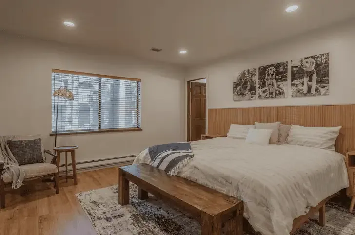 A cozy bedroom with a large bed, wooden bench, armchair, window with blinds, and three dog-themed pictures on the wall above the bed.
