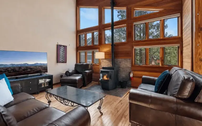 A cozy living room with large windows, leather sofas, a coffee table, a television, a wood stove with fire, and wooden walls and floors, surrounded by forest views.