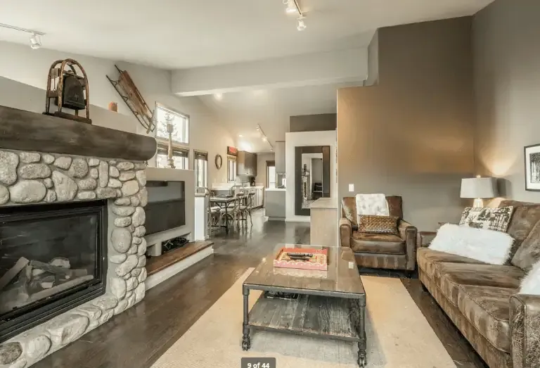 Living room with stone fireplace, leather sofa, armchair, coffee table, and view into dining area; neutral tones and wooden flooring throughout.