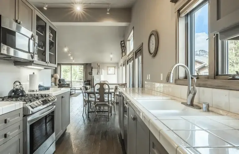 Modern kitchen with stainless steel appliances, light gray cabinets, white tile countertops, large windows, and a dining area in the background with wooden flooring.
