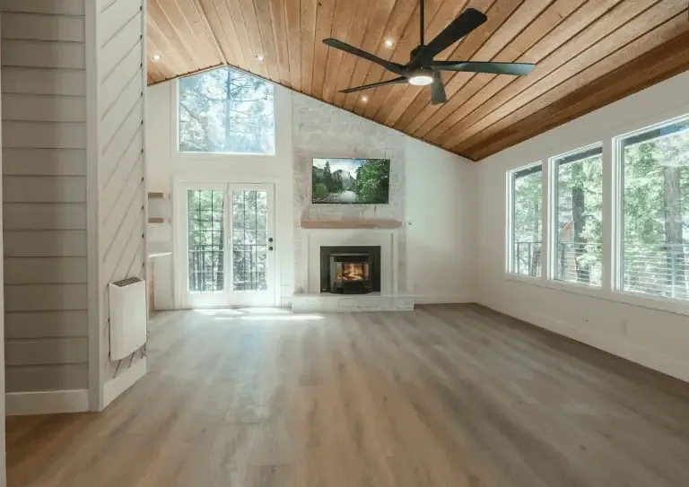 Modern living room with wood-paneled ceiling, large windows, fireplace, mounted TV, and double glass doors opening to a balcony with views of trees outside.